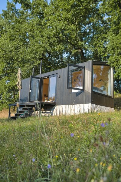 A modern modular cabin with large windows and a small deck sits in a lush meadow surrounded by trees, showcasing sustainable design in a natural retreat setting