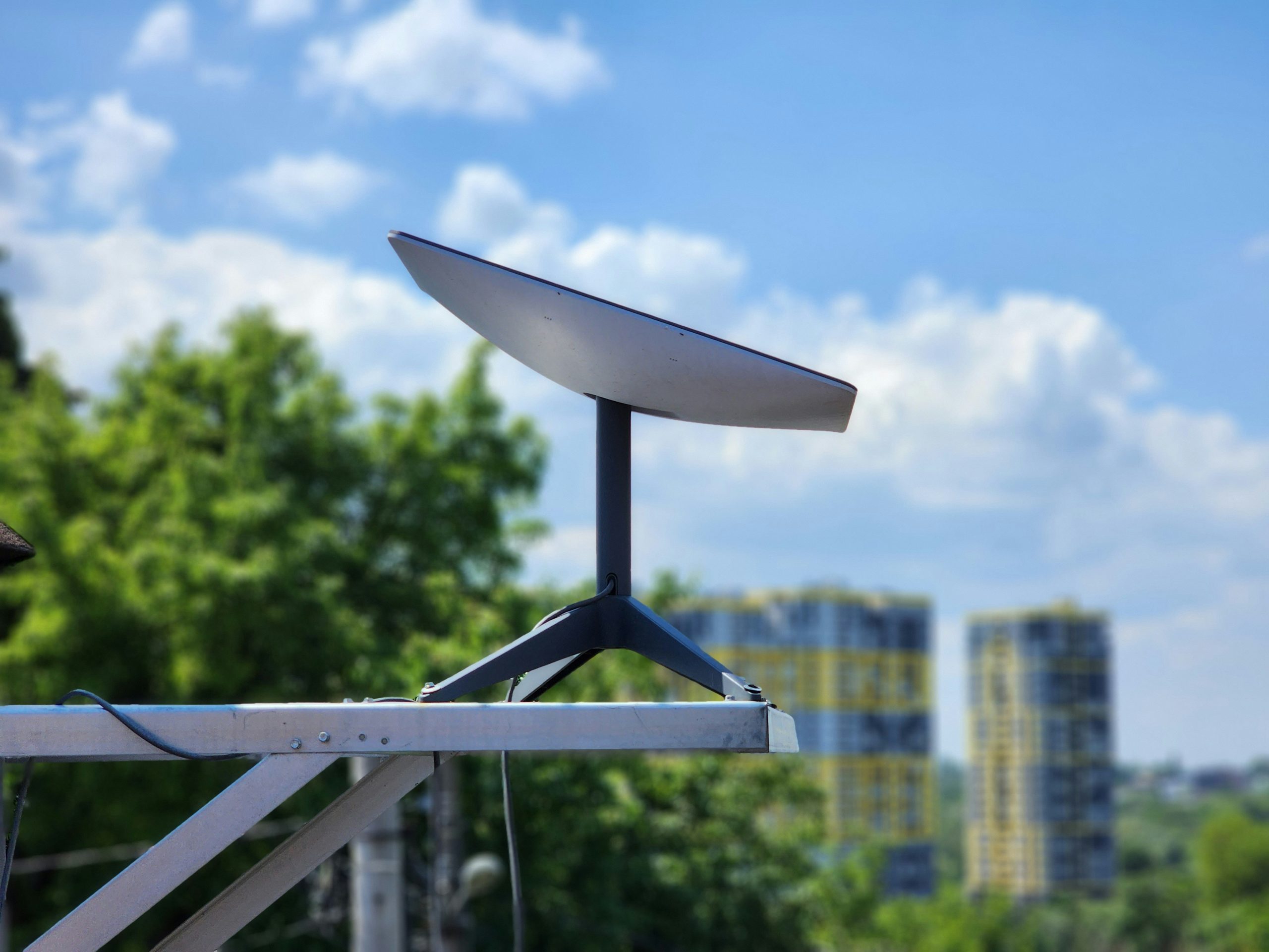 A satellite dish installed on a modular metal frame in an urban setting, with tall buildings and greenery in the background under a clear blue sky