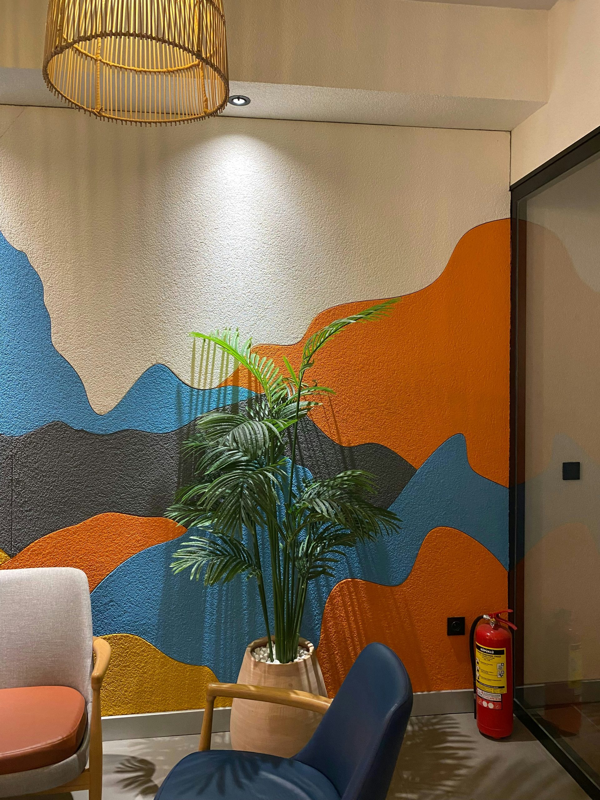 A vibrant office corner featuring a colorful wall with abstract blue, orange, and yellow wave patterns, a potted green plant, modern chairs, and a woven pendant light