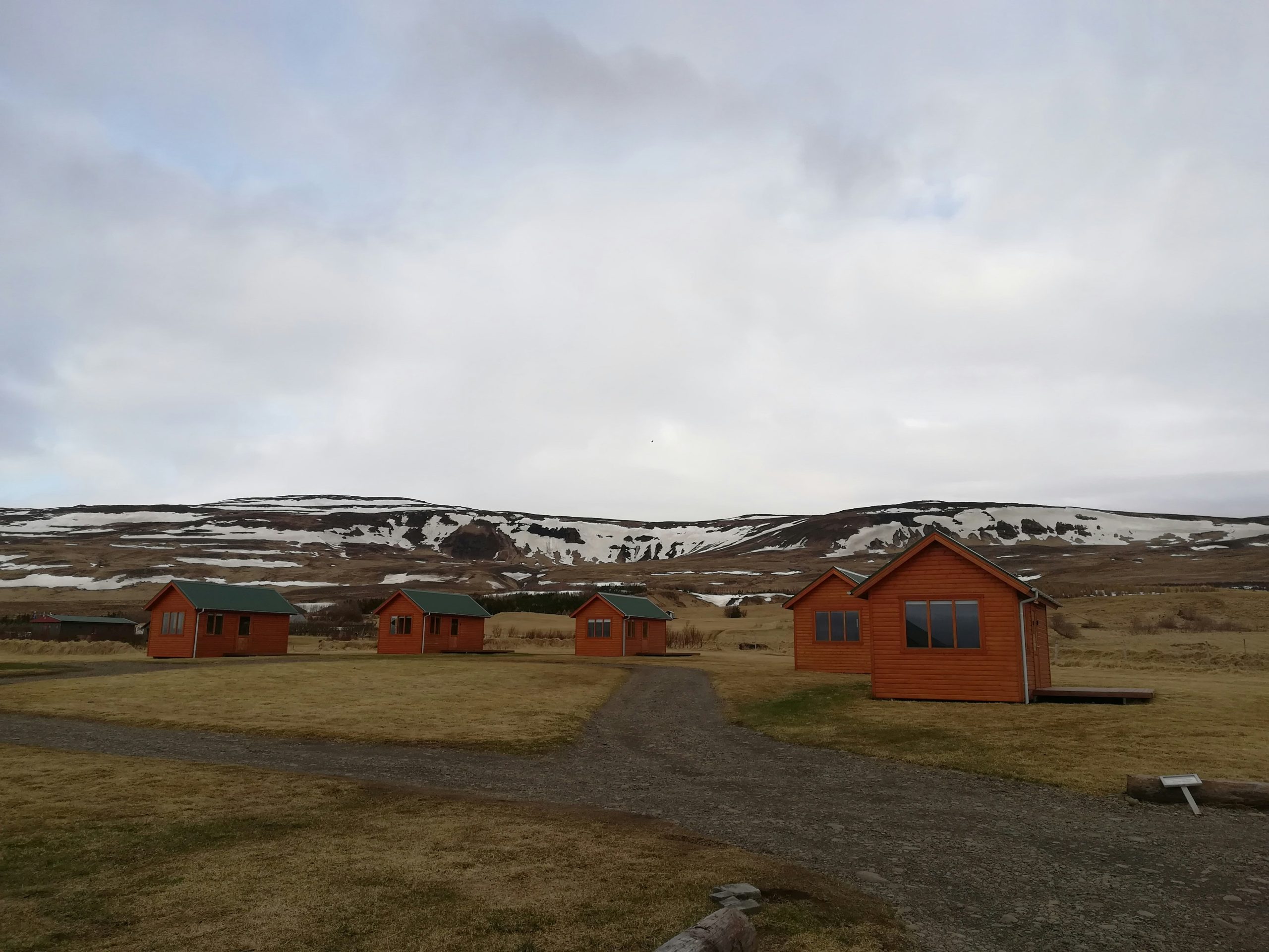 A cluster of orange modular cottages with green roofs sits on a grassy plain, surrounded by a gravel path and distant snow-capped hills under a cloudy sky, ideal for remote modular living