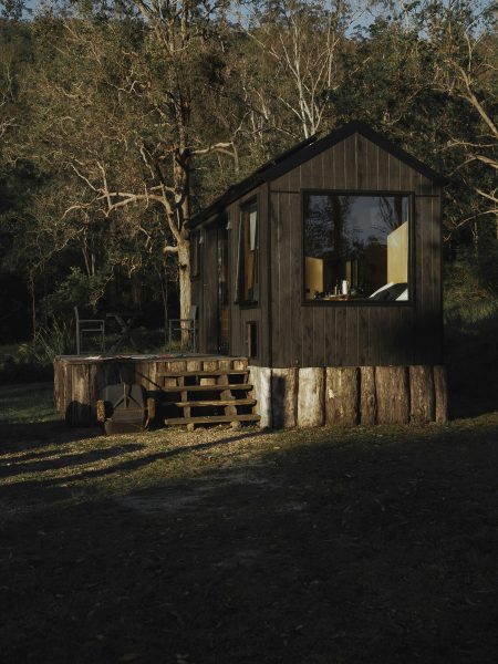 A minimalist modular cabin with dark wood exterior sits in a remote forest clearing, elevated on log stumps with a small wooden staircase leading to a cozy porch area. Surrounded by trees, the cabin features a large window showcasing a warm interior, perfect for off-grid living