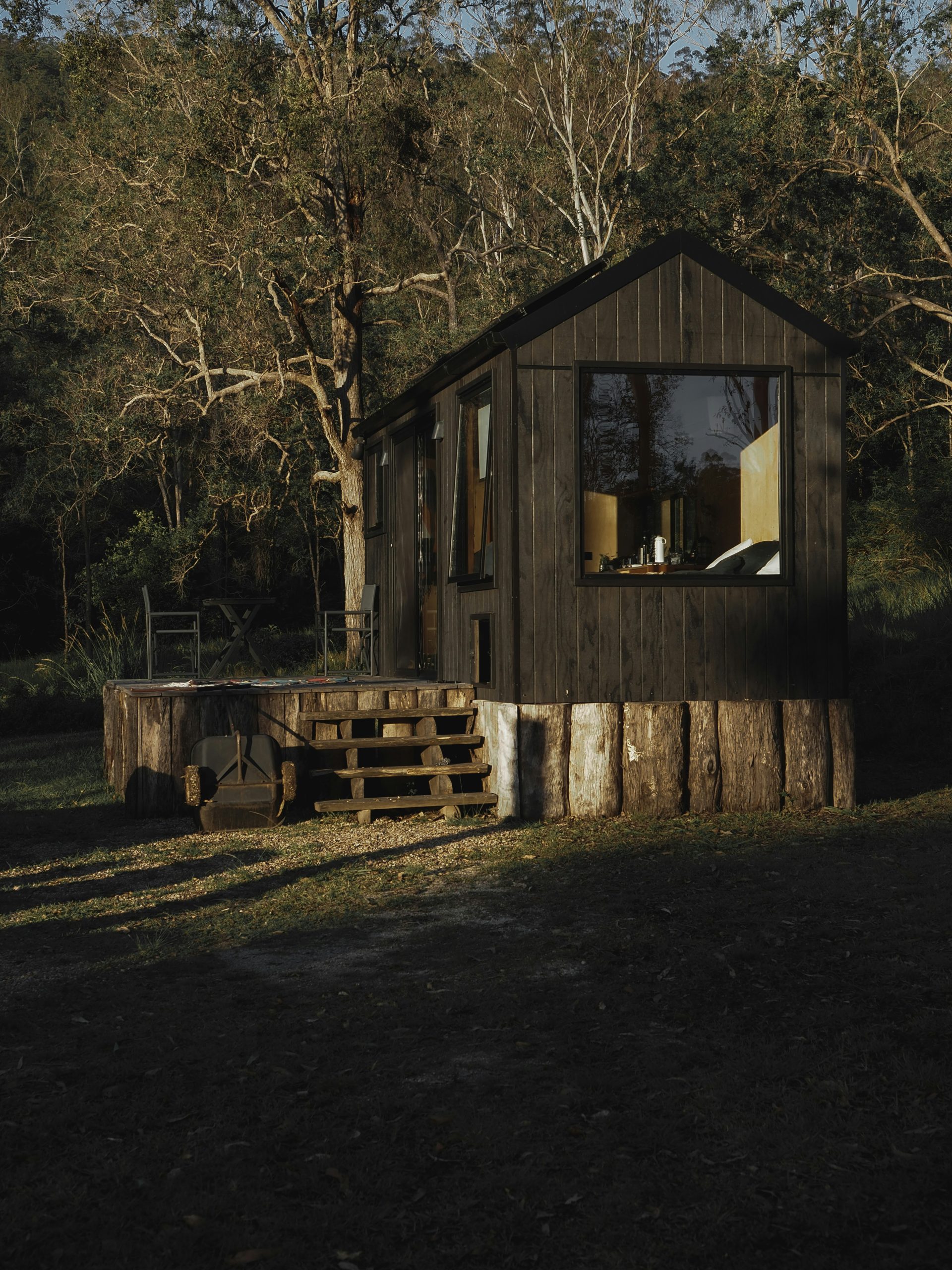 A minimalist modular cabin with dark wood exterior sits in a remote forest clearing, elevated on log stumps with a small wooden staircase leading to a cozy porch area. Surrounded by trees, the cabin features a large window showcasing a warm interior, perfect for off-grid living