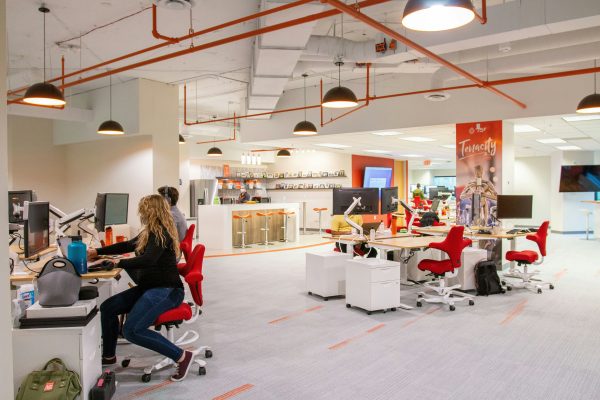 A vibrant open-plan office with employees working at desks with red chairs, featuring modern pendant lighting, orange pipes on the ceiling, and a branded "Tenacity" wall banner in the background