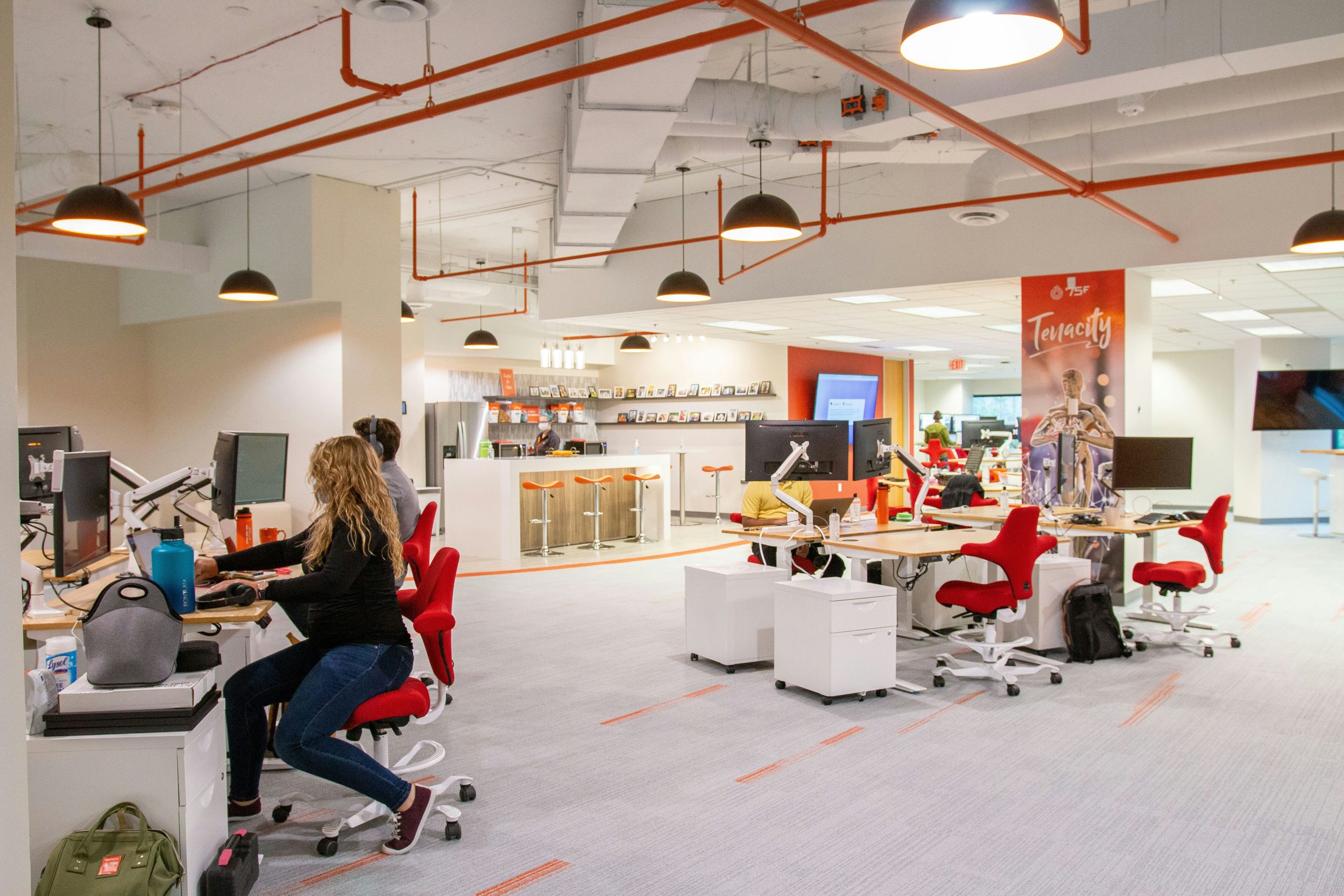 A vibrant open-plan office with employees working at desks with red chairs, featuring modern pendant lighting, orange pipes on the ceiling, and a branded "Tenacity" wall banner in the background