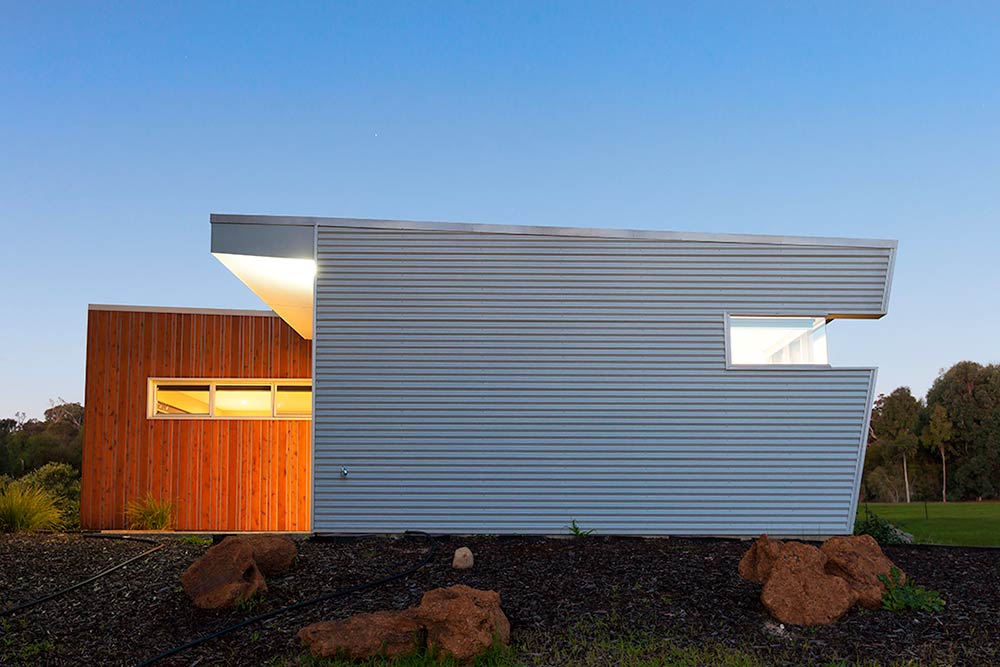 Exterior view of a modular prefab home at dusk featuring corrugated metal siding, warm wooden panels, and natural landscaping